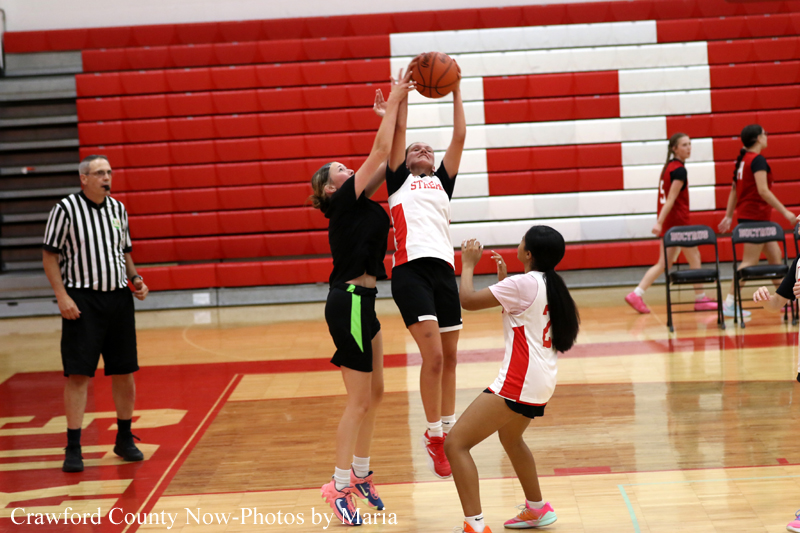 Girls' basketball game: two players leap for the ball in a jump, with a third defender nearby as a referee watches on the left. Red gym bleachers in the background.