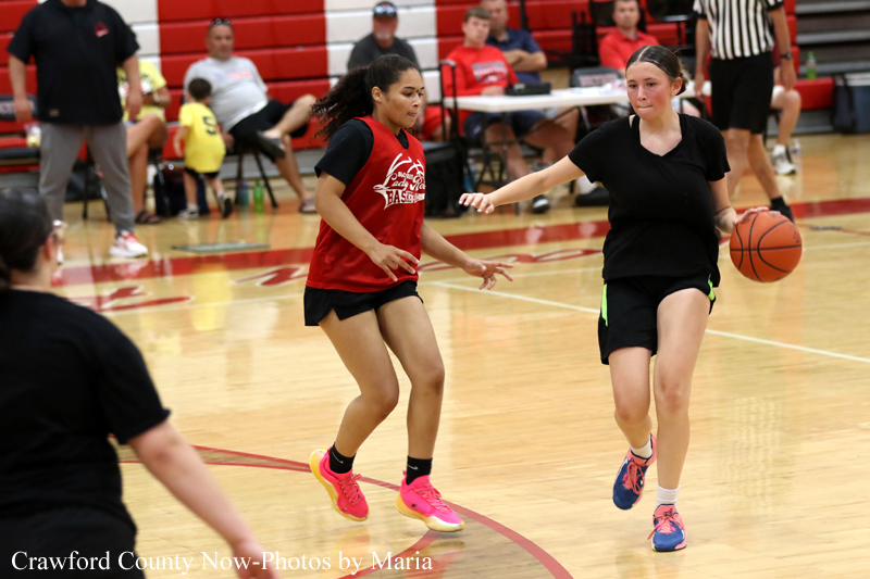 Two young female players compete on a basketball court during a game; one in a red jersey dribbles past a defender in black, with spectators in the background.
