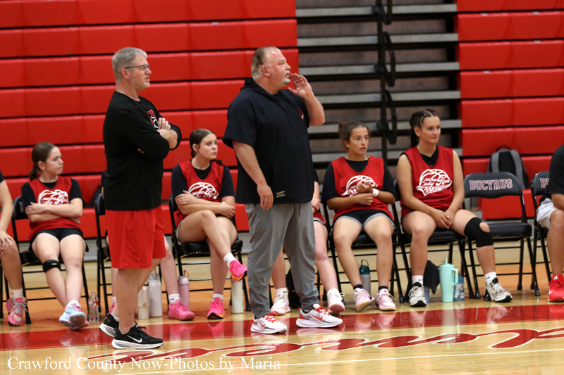 Coaches standing in front of a bench with players seated along a red bleacher wall in a gymnasium during a basketball practice.