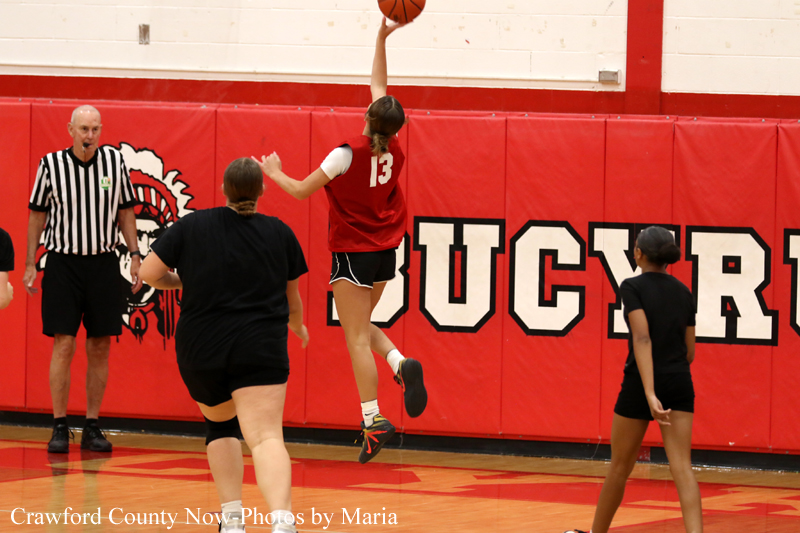 Girl's basketball game: player in red jersey #13 leaps for a shot while a defender and referee watch from the court with red padded walls in the background.