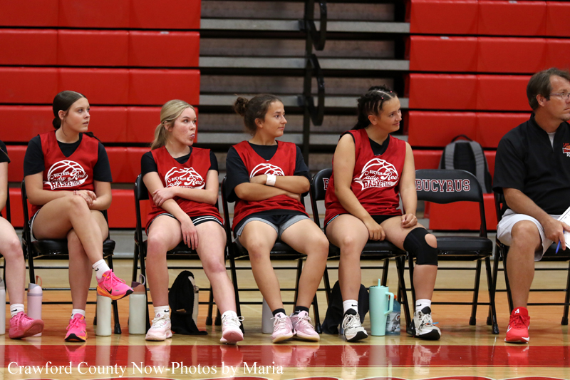 Girls basketball players in red jerseys sit on a bench in a gym, waiting during a game.