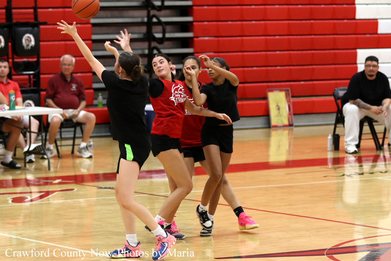 Four young female basketball players reach for the ball during a youth game on a polished gym floor, red bleachers in background.