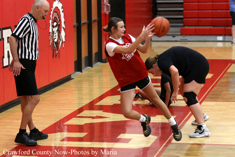 Female player in red jersey trips or falls on a basketball court as a referee watches nearby on a wood floor with red padding on walls behind.
