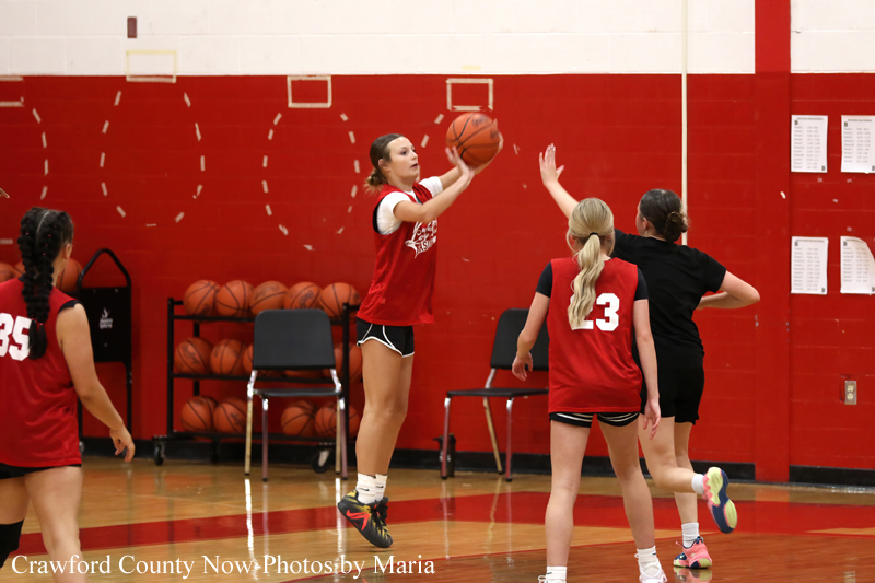 Young female basketball player in a red jersey jumps to shoot a ball during a drill as teammates defend in a gym with red walls.