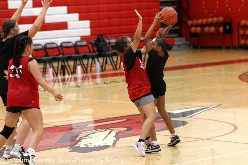 Girls' basketball game in a gym; players in red jerseys guard and contest the ball near the key while a defender in black reaches for a pass.