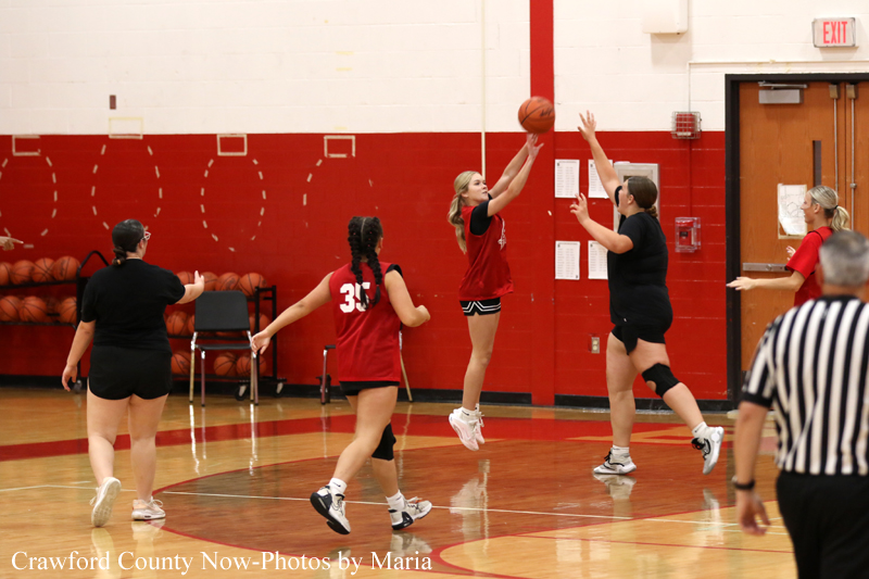 Girls' basketball game in a gym: player in red jumps to shoot while defender reaches up to block, with teammates and a referee nearby.
