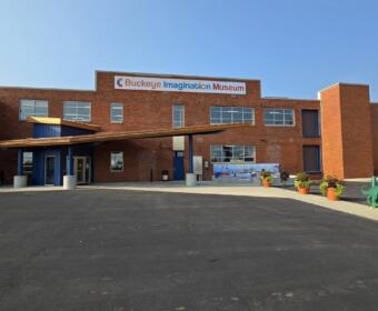 Brick exterior of Buckeye Imagination Museum with a blue entry canopy and potted plants outside the entrance.