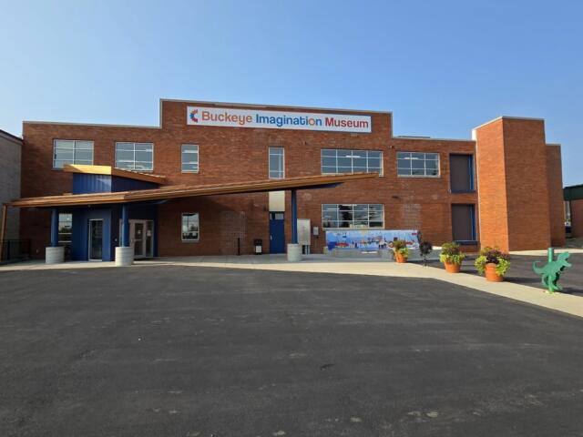 Brick exterior of Buckeye Imagination Museum with a blue entry canopy and potted plants outside the entrance.