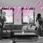 Two cleaners in overalls vacuuming and mopping a modern living room beneath a pink neon sign that reads 'Cleaning by Carrie'