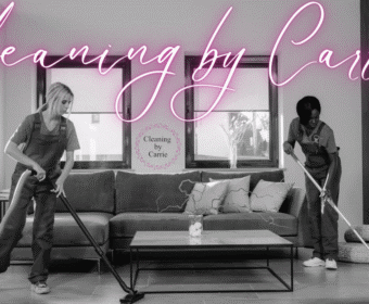 Two cleaners in overalls vacuuming and mopping a modern living room beneath a pink neon sign that reads 'Cleaning by Carrie'