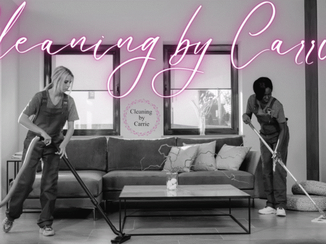 Two cleaners in overalls vacuuming and mopping a modern living room beneath a pink neon sign that reads 'Cleaning by Carrie'