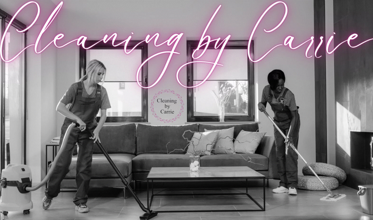 Two cleaners in overalls vacuuming and mopping a modern living room beneath a pink neon sign that reads 'Cleaning by Carrie'
