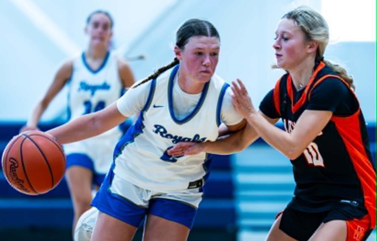 Two female basketball players compete for the ball during a fast dribble, one in white jersey and the other in black-red defense in a gym.