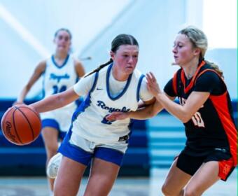 Two female basketball players compete for the ball during a fast dribble, one in white jersey and the other in black-red defense in a gym.