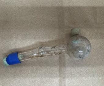 Dirty glass laboratory tube with a round bulb at one end and blue stopper on the other, lying on a bare concrete surface (informational).