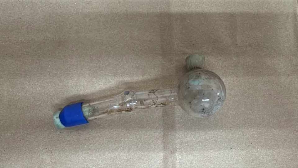 Dirty glass laboratory tube with a round bulb at one end and blue stopper on the other, lying on a bare concrete surface (informational).