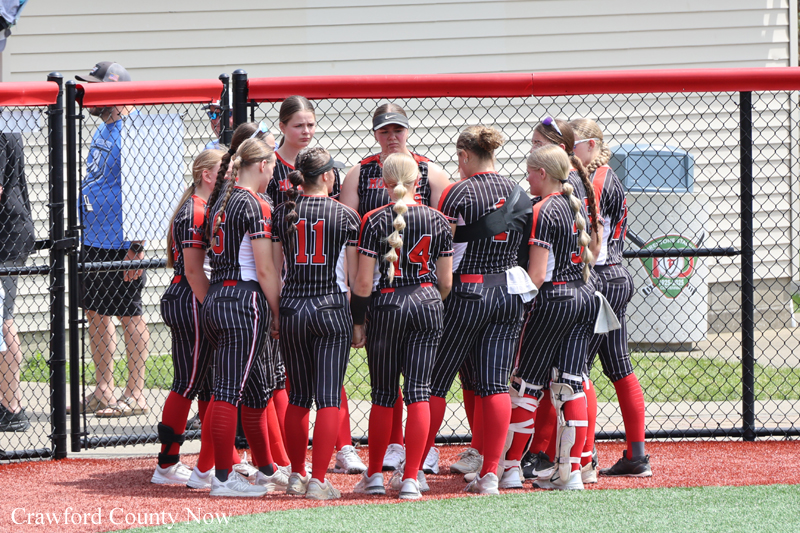 Softball team huddle on the field in black pinstripe uniforms with red numbers and red socks, gathered in a circle near a chain-link fence.