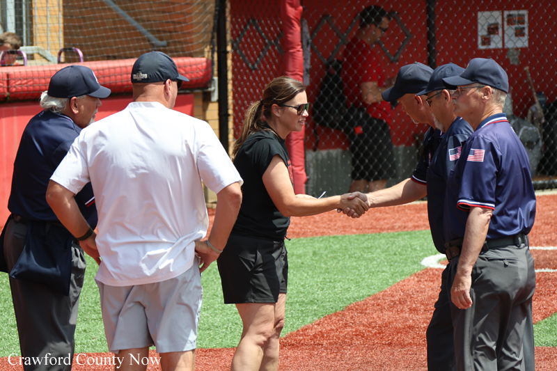 Woman in a black shirt shakes hands with an umpire in navy uniform while others watch on a baseball field.