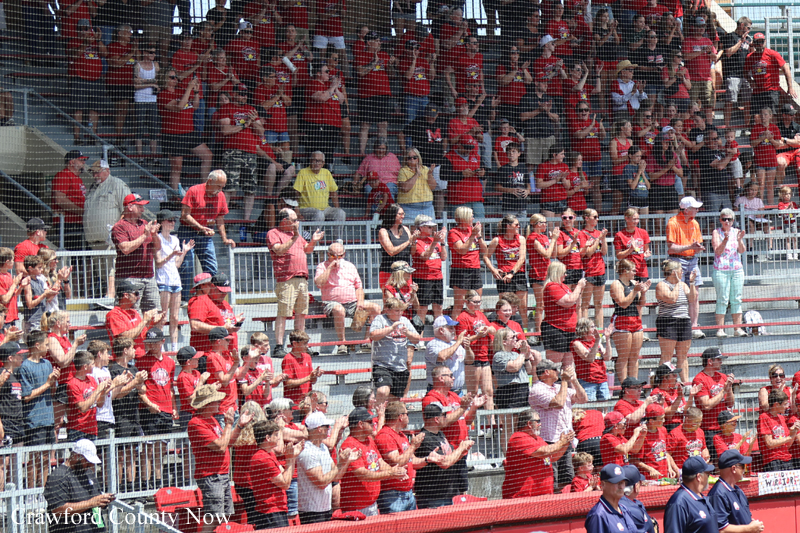 Large crowd of fans in red clothing standing and clapping behind a safety net at a sports stadium stands.
