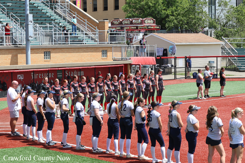Two rows of girls in blue-and-white uniforms stand on a track at Firestone Stadium for a ceremony, with bleachers in the background.