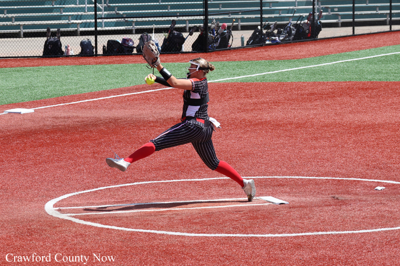 Softball pitcher mid-pitch on a red dirt mound, wearing pinstripe uniform, red socks, and glove with a green-felt ball in hand, bleachers in background.
