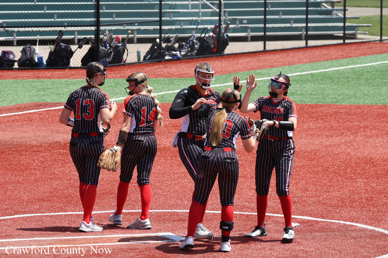 Girls' softball team in black pinstripe uniforms with red numbers high-fiving on the red infield near home plate, with bleachers in the background.