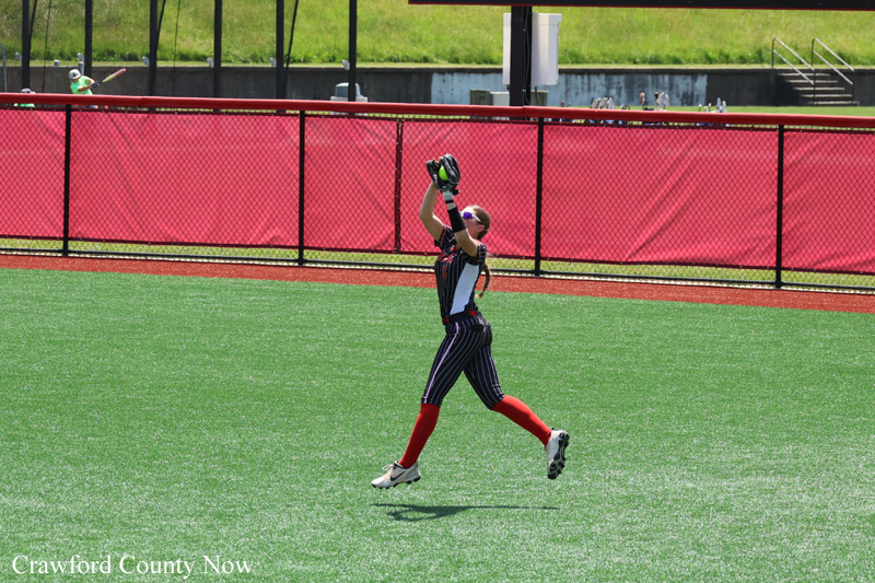 Female softball player in pinstriped uniform jumps to catch with glove raised above her head on a green field, red outfield fence behind her.