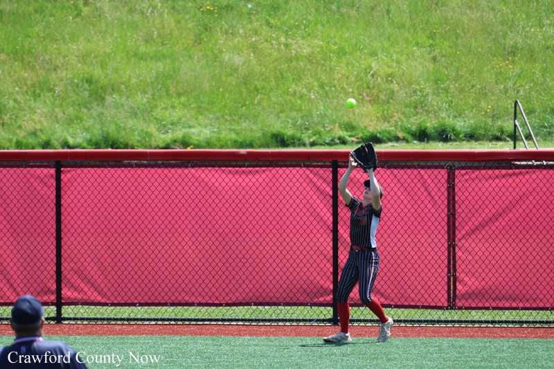 Softball fielder in a striped uniform raises glove to catch a ball near a red outfield fence on a grassy field.