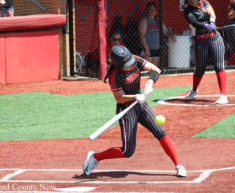 Female softball player in black-and-red striped uniform swings a bat at a pitched ball on a red dirt field.