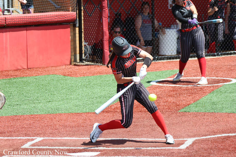 Female softball player in black-and-red striped uniform swings a bat at a pitched ball on a red dirt field.