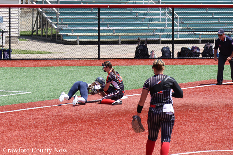 Softball game moment: baserunner slides into a base as a catcher and teammate reach to tag, on a red infield with empty bleachers in the background.