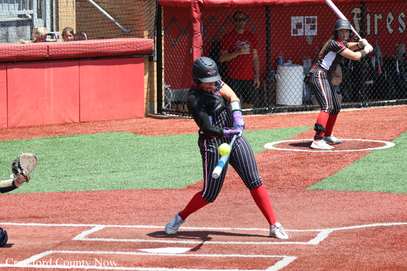 Young female softball player in a black pinstriped uniform swings a bat at a pitched ball on a red-dirt field, wearing a helmet and red socks; another player stands ready in the background near the dugout.