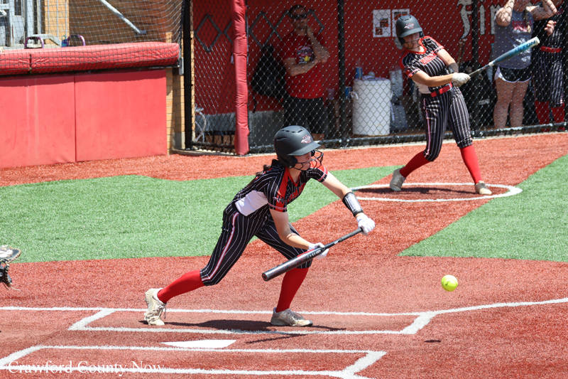 Young softball player in a red-and-black uniform swings at the plate with a helmet on, near a teammate and chain-link fence as spectators watch.
