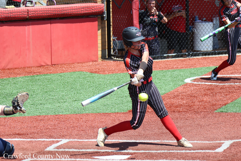A female softball player in a pinstriped uniform swings at a pitched ball, wearing a helmet and red socks on a dirt and artificial turf field, with a catcher’s glove visible on the left edge.