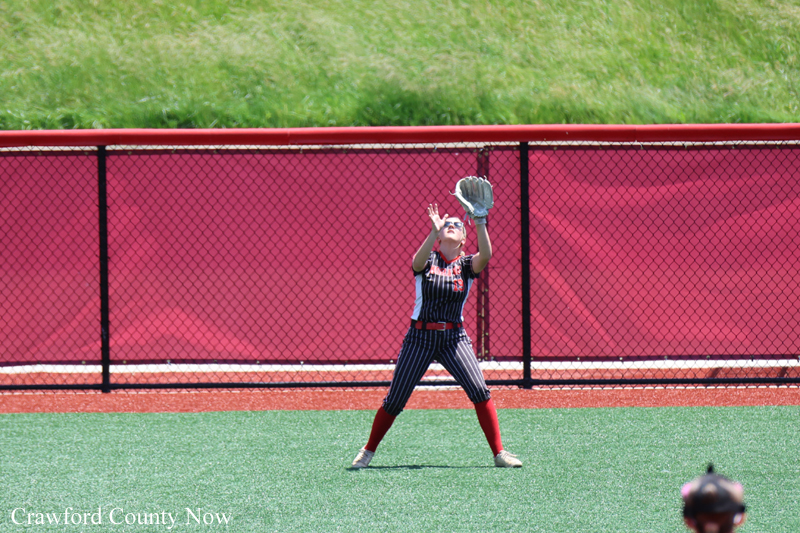 Young baseball/softball fielder in pinstripe uniform reaches up with a glove to catch a ball on a green field with a red fence behind him.