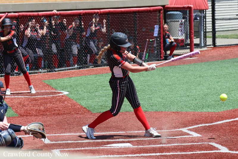 A young female softball batter in a black pinstripe uniform swings at a pitch during a game, with cheering fans in the dugout area behind."