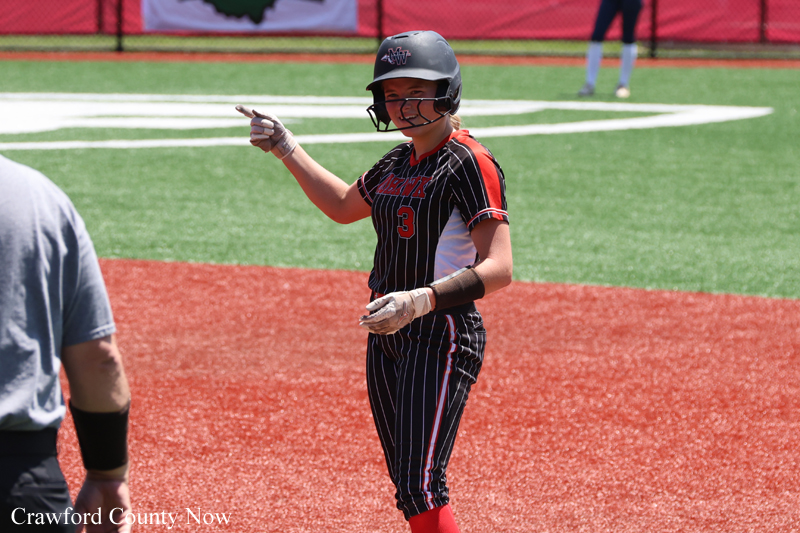 Baseball player in a black pinstripe uniform with red trim and number 3 points with their gloved left hand while wearing a helmet on a sunny baseball field.