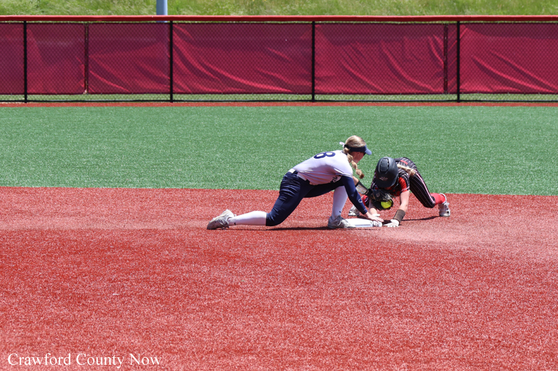 Two softball players slide into a base during a tag play on a red dirt infield, with a defender reaching for the tag against a green outfield backdrop.