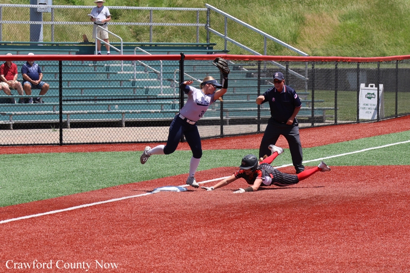 Softball play at home plate: a runner slides in as a fielder jumps to tag, with an umpire watching nearby on the infield.