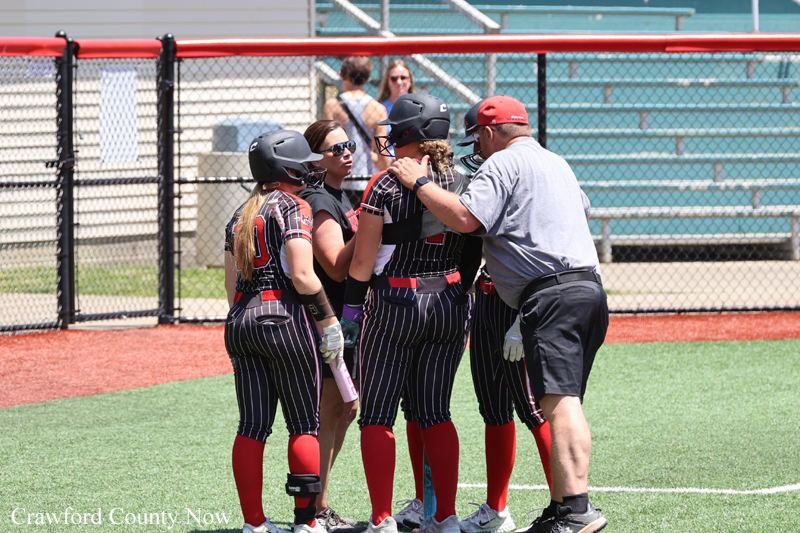 Softball team huddle on the field as coach gives guidance to players wearing helmets and striped uniforms.