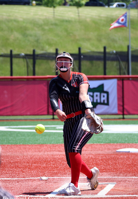 Softball pitcher in a black pinstripe uniform mid-pitch on the mound, wearing a face-guard helmet and red socks.