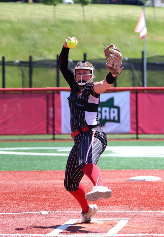 Female softball pitcher mid-throw on the mound, wearing a helmet with face mask and glove, in a pinstriped uniform.