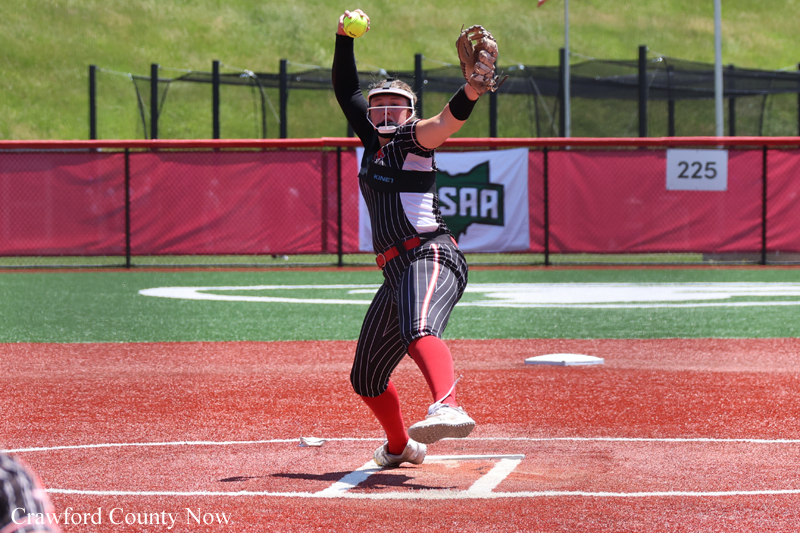 Softball pitcher in a black pinstripe uniform winds up to throw on a red infield with green grass beyond, glove raised.