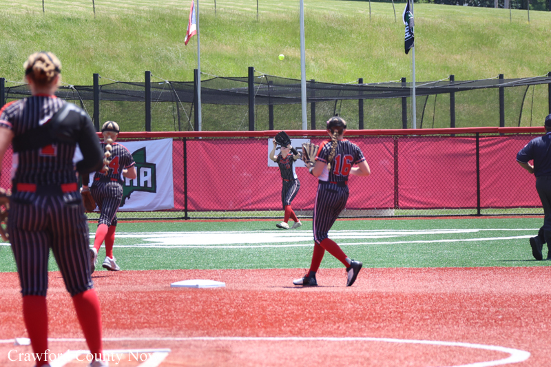 Young softball players in black-and-red pinstriped uniforms play on a bright field, with a pitcher mid-throw and fielders ready in the infield/outfield fence area.