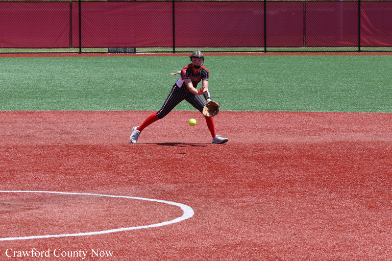 Softball infielder in red and black uniform fields a ground ball on a red infield with a green outfield in the background (Crawford County Now watermark).