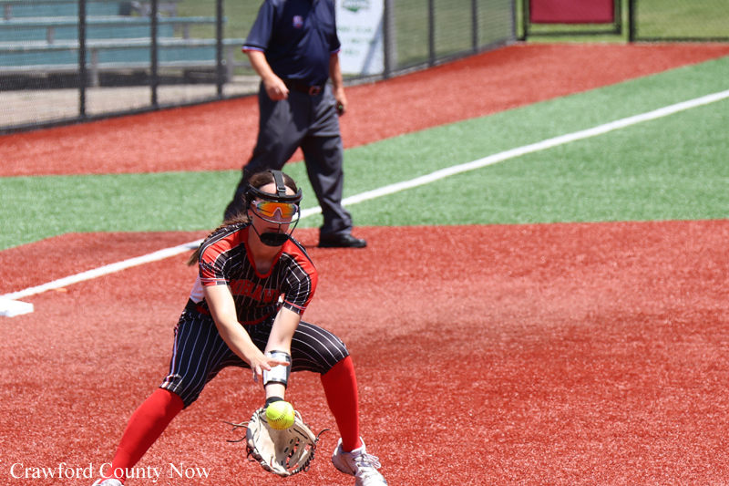 Female softball catcher in red-and-black uniform crouches to field a ground ball on the infield, wearing a protective mask and glove with red socks visible. Umpire stands in the background.