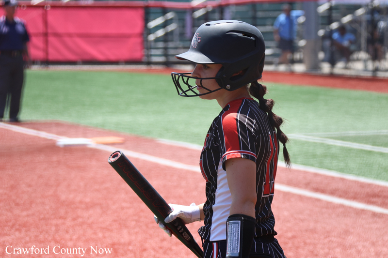 Young female softball batter in helmet and striped uniform, gripping a bat at the on-deck circle on a sunny field