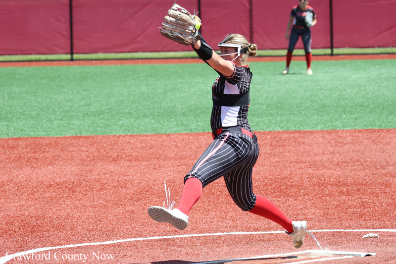 Softball pitcher in a black pinstripe uniform delivering a throw from the mound on a red dirt infield, green outfield behind her.