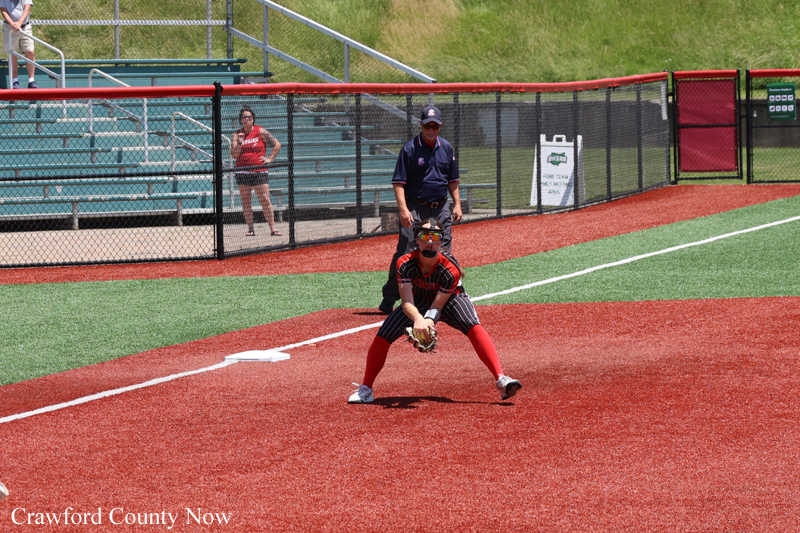 Softball fielder in red and black crouches to field a ground ball on a red infield, with an umpire and a spectator behind the fence.