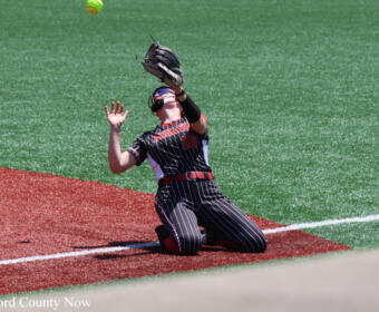 Softball fielder in pinstriped uniform dives on red dirt, glove raised to catch an airborne ball on a green field nearby.
